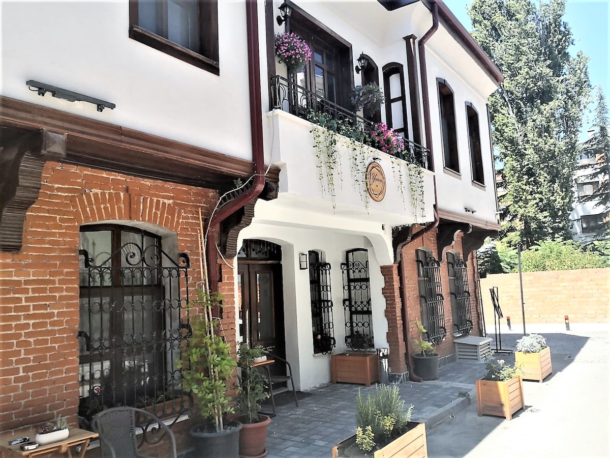 The exterior of the hotel is characterized by a blend of exposed brick and white walls, accented by decorative ironwork on the windows. Potted plants and flowers adorn the entrance, providing a touch of greenery against the backdrop of a sunny day.