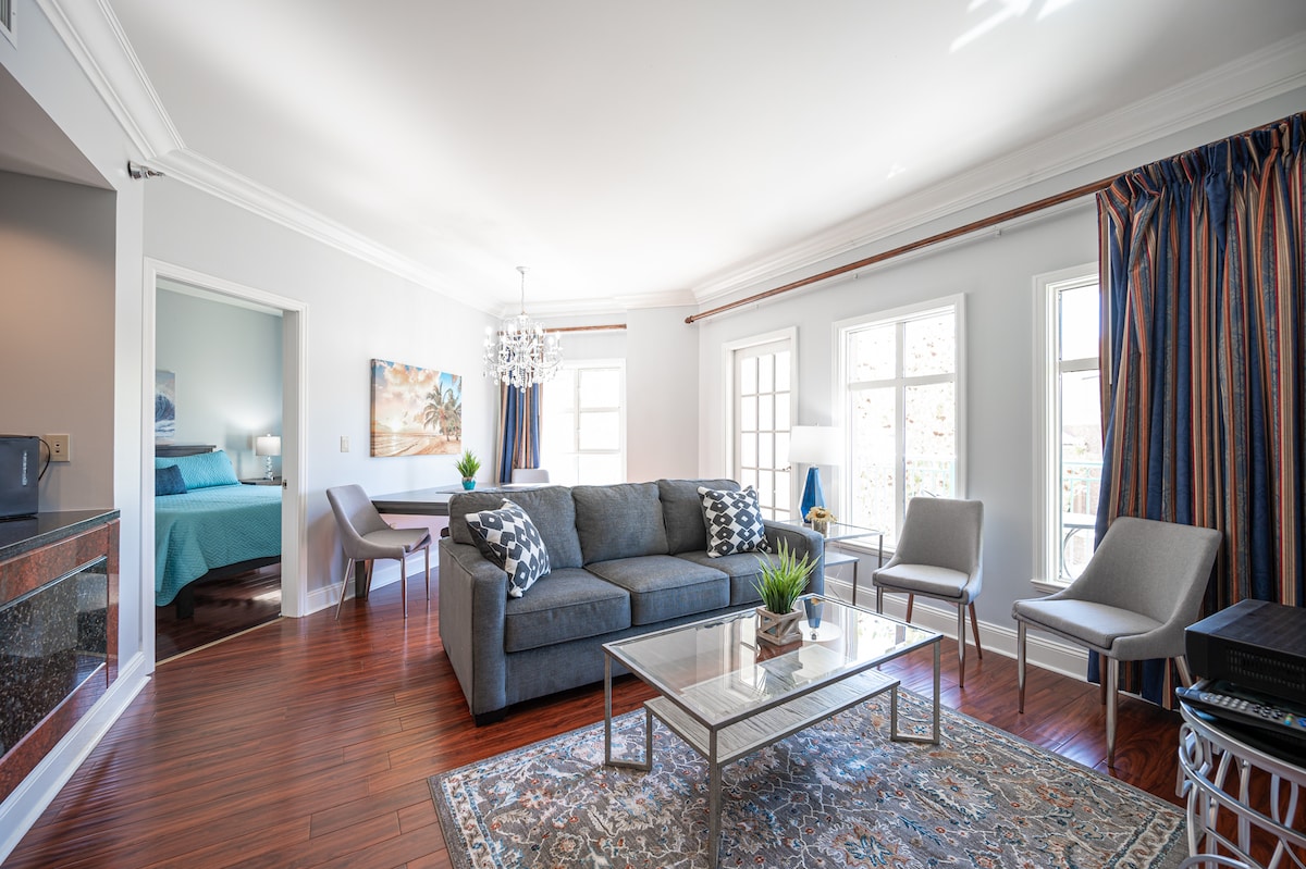 A spacious living area is presented, featuring a comfortable gray sofa with decorative pillows. A glass coffee table sits atop an area rug, complemented by four stylish chairs. Large windows allow natural light to fill the room, offering views of the outside landscape.