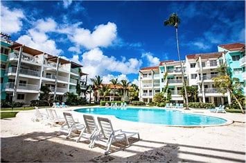 A vibrant pool scene is captured, featuring a large, inviting swimming pool surrounded by well-kept green lawns. A cluster of lounge chairs is arranged near the pool, with palm trees adding to the tropical atmosphere. Spacious apartment buildings provide a colorful backdrop.