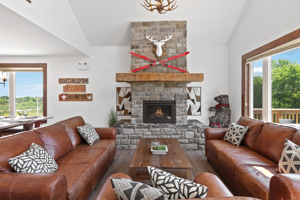 A spacious living area showcases two large leather sofas, arranged around a coffee table. A stone fireplace, adorned with rustic decor including antlers and skis, serves as a focal point. Large windows allow natural light to flood the room, offering views of the outdoors.