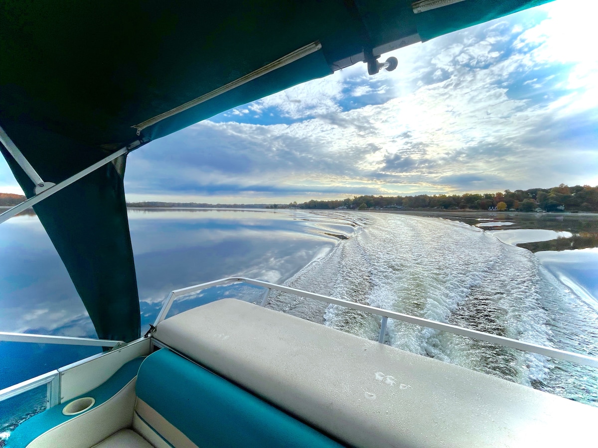 The image captures the view from the rear of a pontoon boat, showcasing a tranquil lake at sunrise. Gentle ripples are seen in the water, reflecting hues of blue and gray in the sky, while a soft wake trails behind the boat.