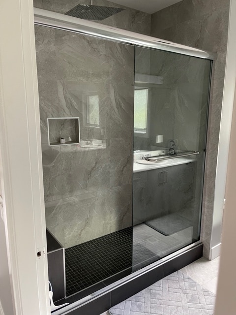 A modern bathroom is featured with a glass-enclosed shower, showcasing sleek, gray marble walls. A minimalist sink area is visible in the background, with ample natural light illuminating the space. A textured bath mat is positioned on the floor for comfort.