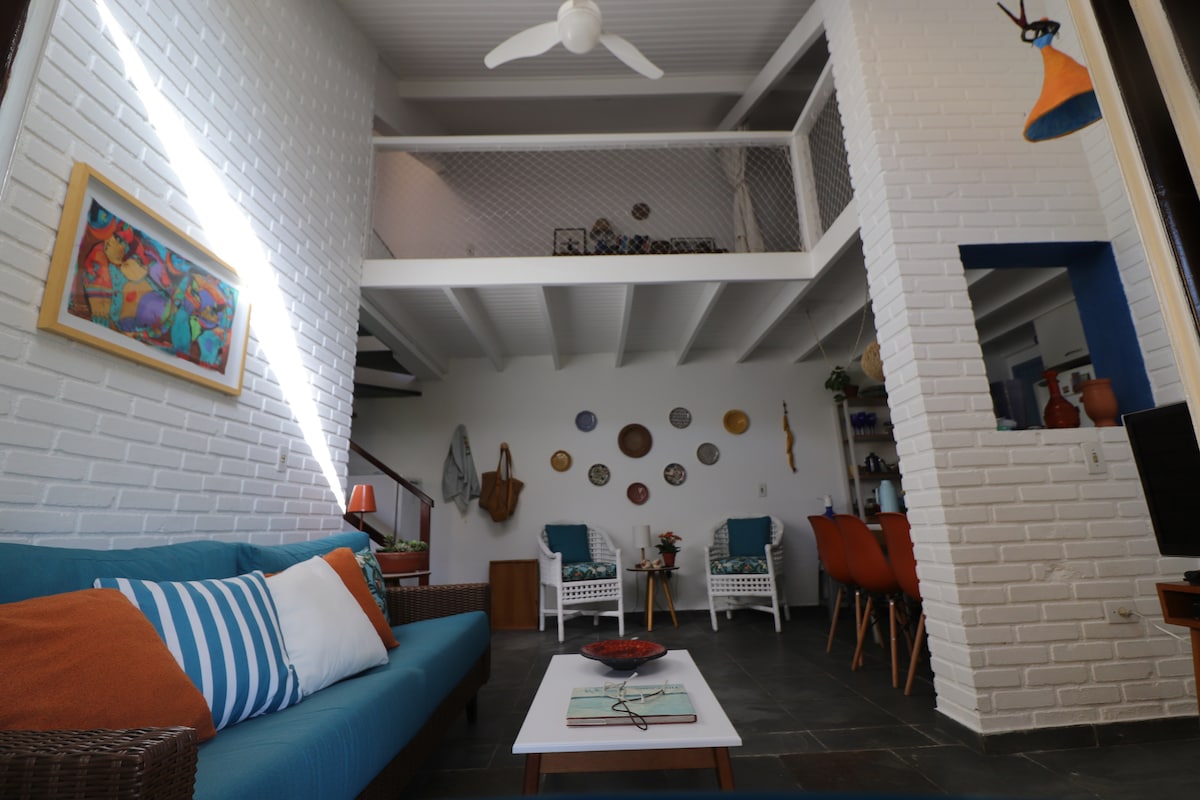 A bright living space is offered, featuring a comfortable blue sofa with decorative pillows. The wall is adorned with colorful artwork and plates. Above, the mezzanine adds depth, while a dining area with orange chairs is visible in the background. Natural light enhances the openness.