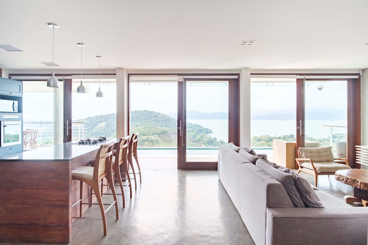 An open-concept living area is showcased, featuring large glass doors that offer a scenic view of the surrounding hills and bay. Natural light fills the space, which includes a modern kitchen and a spacious seating arrangement with a soft gray sofa. Wooden accents are present throughout.