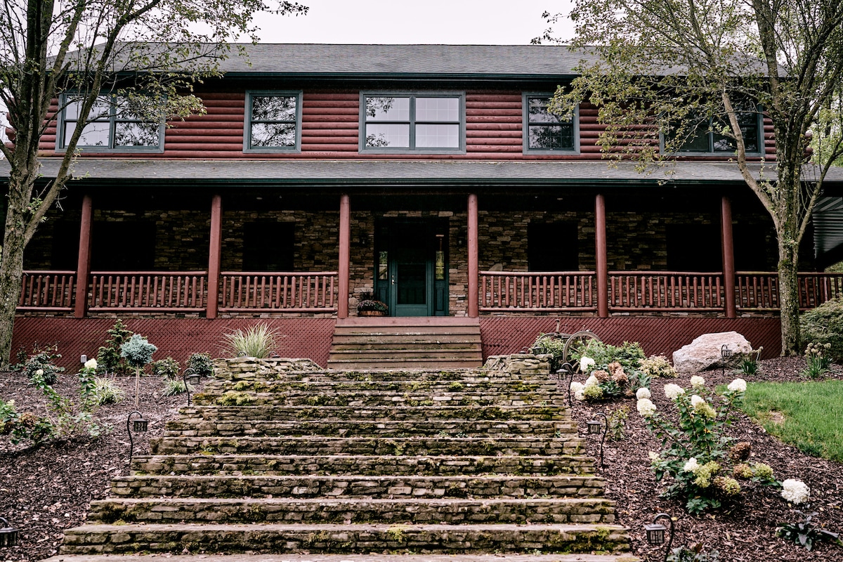The entrance of a large, two-story house features a stone facade and wooden accents. A set of wide stone steps leads up to a covered porch, framed by tall support columns. Surrounding gardens include lush greenery and blooming flowers.