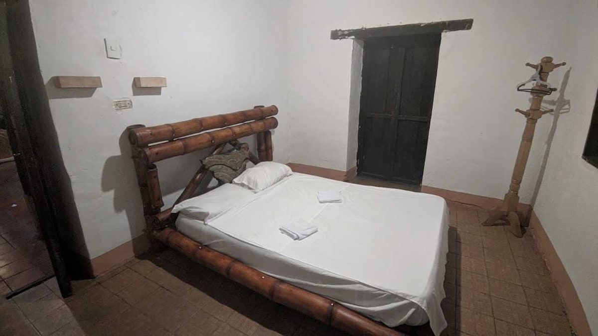 A simple and cozy sleeping area features a bamboo bed with a white mattress and towels neatly arranged. The light-colored walls contrast with a dark wooden door and a wooden coat rack, creating a minimalistic yet welcoming atmosphere.