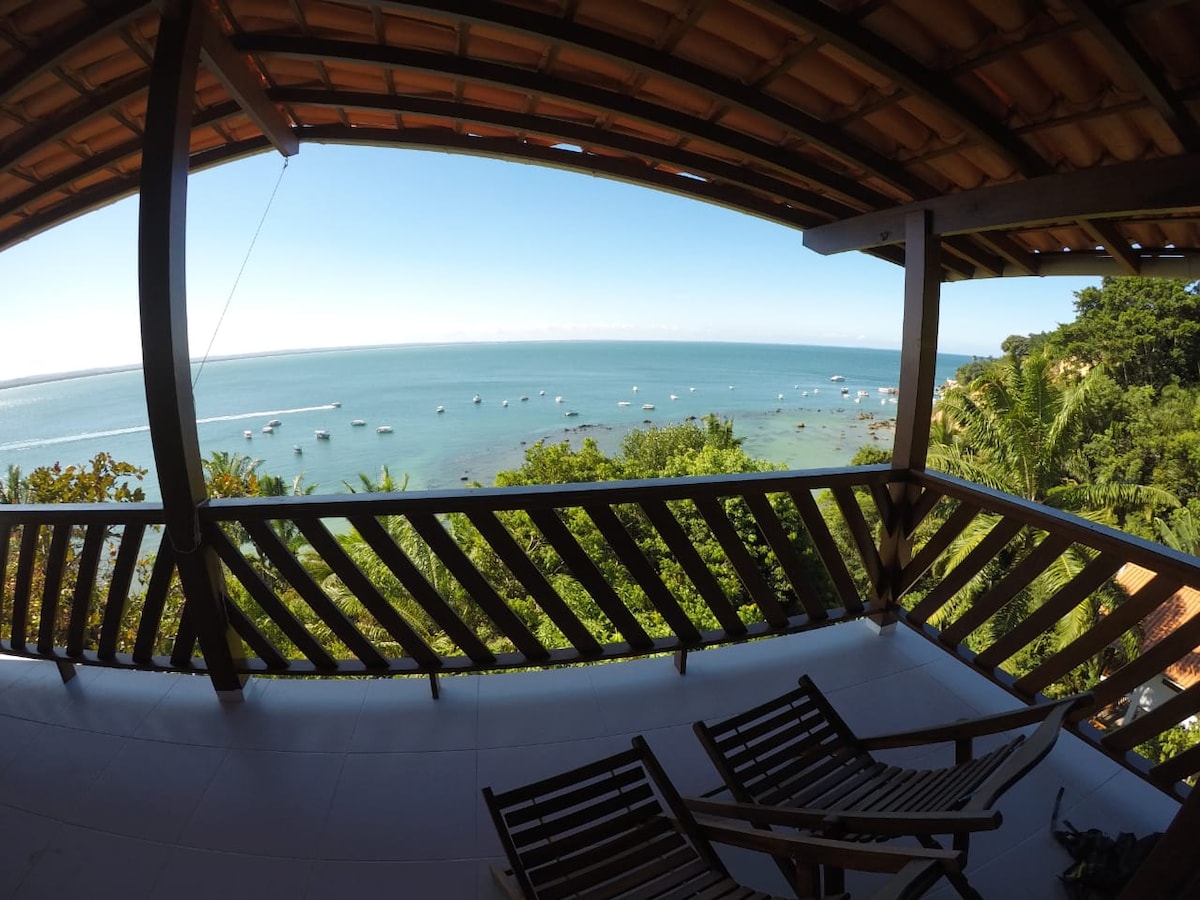 A spacious balcony offers a panoramic view of the sea, framed by wooden railings. Two lounging chairs are positioned to take in the scenery, with lush greenery surrounding the area. The tranquil ocean is dotted with small boats, enhancing the serene atmosphere.