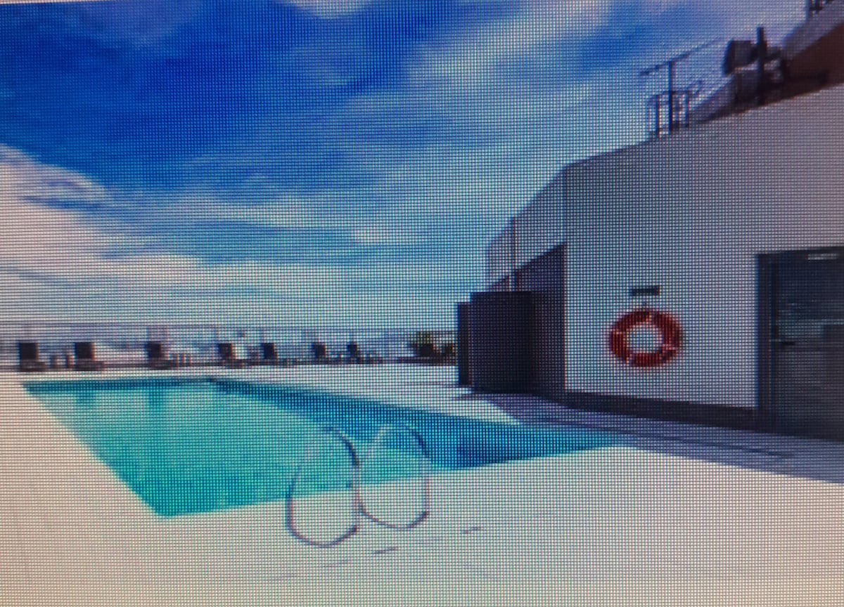 A rooftop pool area is viewed, featuring a clean, rectangular swimming pool surrounded by a spacious deck. Lounge chairs are positioned nearby, and a lifebuoy is mounted on the wall. The sky above is bright, with scattered clouds enhancing the open feel.