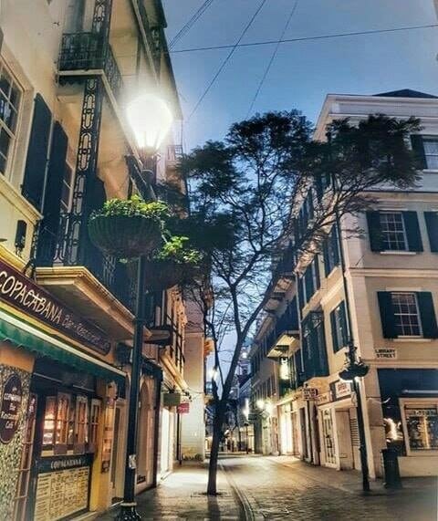 A quiet street scene is captured in soft evening light, featuring traditional architecture with dark green shutters. A streetlamp casts a warm glow, illuminating a small tree alongside the cobbled pathway. Shops and cafes line the street, creating an inviting atmosphere.