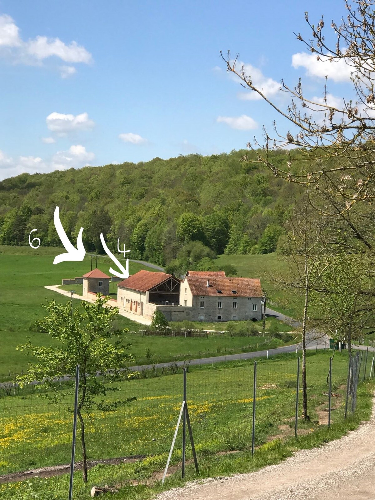 A scenic view of a rustic property set against a backdrop of lush green hills and clear blue skies. Two buildings are visible, indicating a welcoming structure for guests. A tranquil pathway leads through the surrounding landscape, enhancing the sense of seclusion and nature.