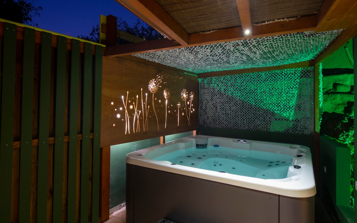A private jacuzzi is set beneath a wooden canopy, surrounded by decorative wooden panels. Soft green lighting enhances the tranquil ambiance, while the wall features an illuminated floral design, contributing to a calming environment for relaxation.
