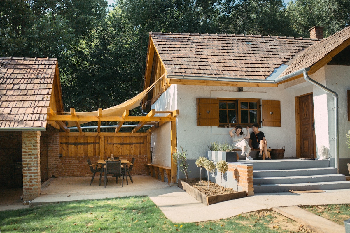 The exterior of a charming white house is showcased, featuring a wooden terrace with chairs and a table. A pergola extends from the house, providing shade. The surrounding greenery adds to the serene atmosphere. Nearby, a rustic structure complements the main house.