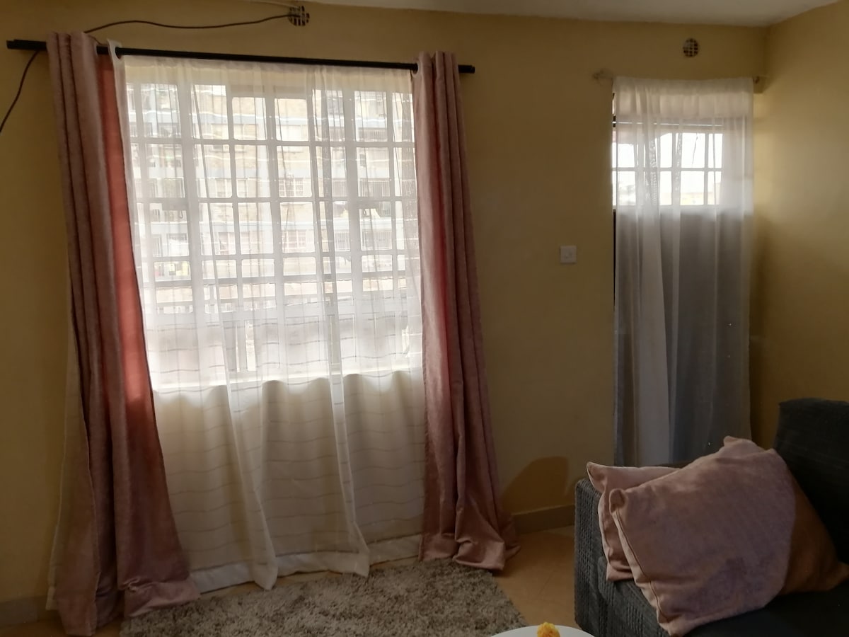 A sunlit living area features large windows draped with sheer and solid curtains in soft hues. A plush rug covers the floor, complementing the neutral wall color. A cozy grey sofa with decorative cushions is positioned nearby, creating a comfortable seating space.