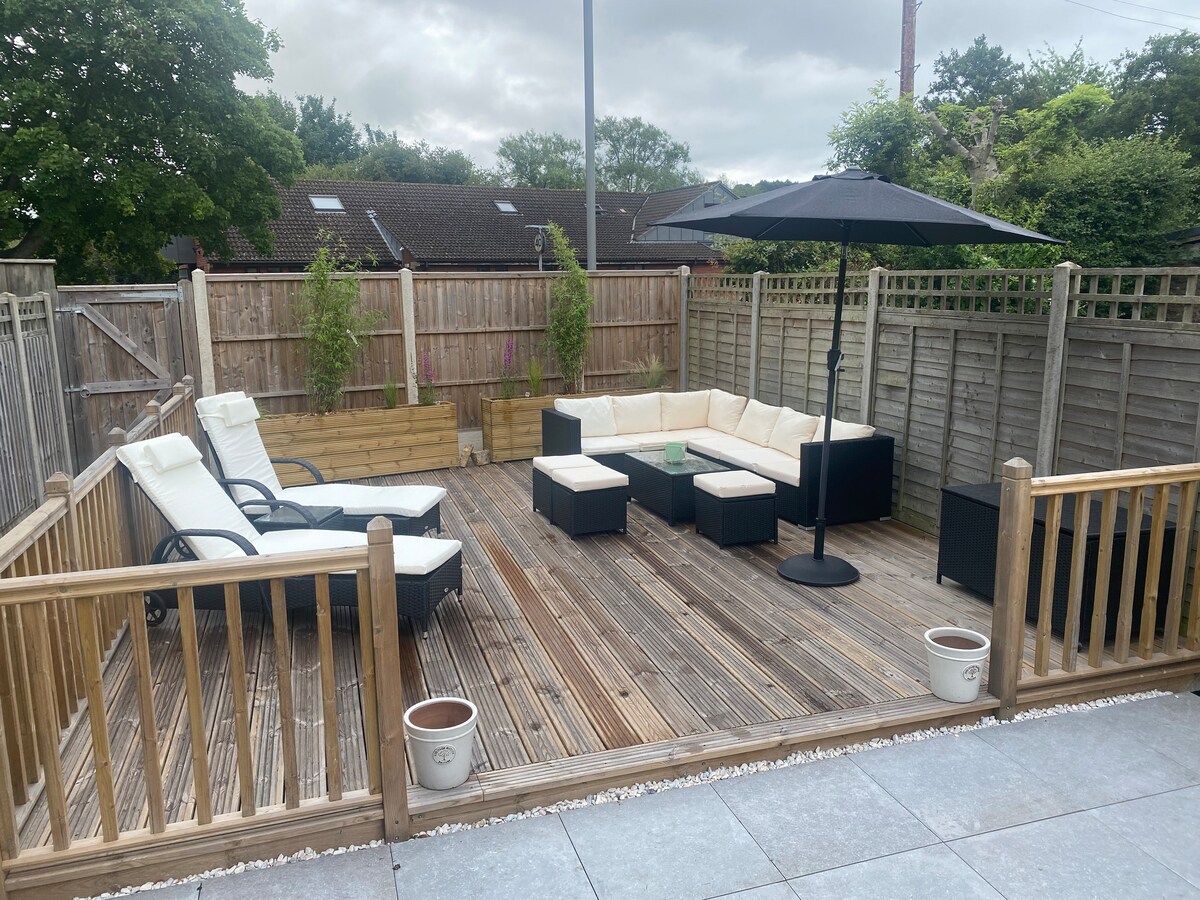 The outdoor deck features a spacious seating area with light-colored cushions on a dark frame. A large umbrella provides shade over the seating arrangement. Sun loungers sit nearby on the wooden surface, surrounded by a fenced garden with greenery in the background.