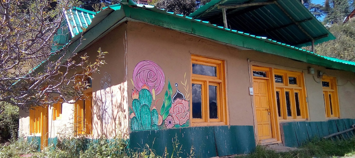 A charming mountain homestay features a vibrant mural on the side, showcasing floral designs. Bright yellow windows frame the structure, contrasting with the earthy tones of the walls. The building is surrounded by greenery, and a sloped roof adds to its rustic appeal.