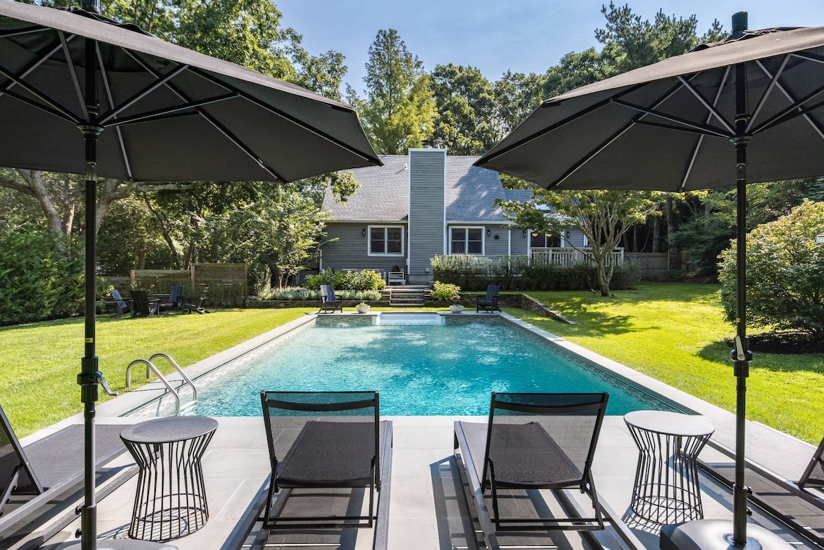 A serene outdoor space featuring a refreshing pool surrounded by lush greenery. Dark lounge chairs and umbrellas offer shade near the pool, which is complemented by the home's subtle, natural tones in the background. The atmosphere is inviting for relaxation and leisure.