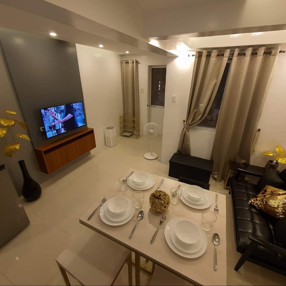 A dining area features a neatly set table with plates and glasses, accompanied by a black leather sofa. Natural light enters through the nearby window, while a television is positioned on the wall across from the seating area. Soft curtains frame the window and enhance the space.