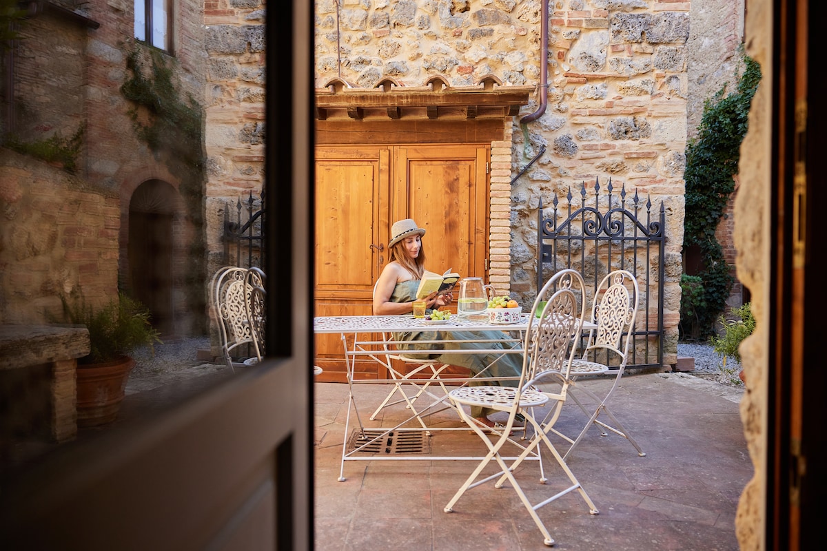An outdoor dining area features a wrought-iron table surrounded by six matching chairs. A wooden sideboard is positioned against a rustic stone wall. Potted plants add greenery to the setting, while a gate frames the scene, enhancing the charming atmosphere.