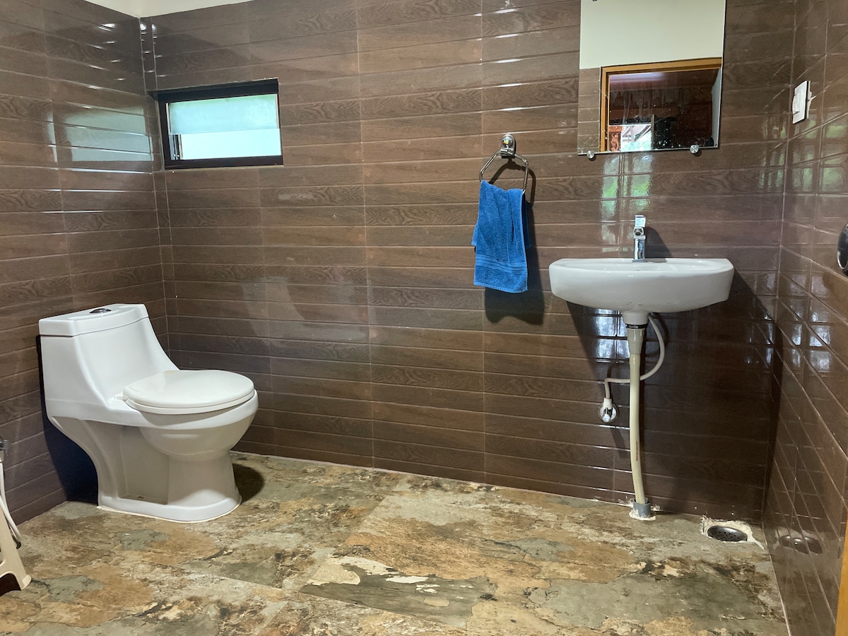 A bathroom features modern fixtures against brown tiled walls. A white toilet and a sink with a chrome faucet are included. A blue towel is hung on a wall-mounted hook, and natural light enters through a small window.