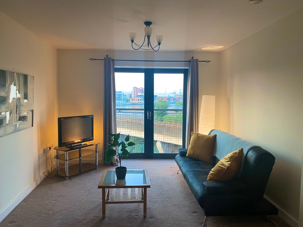 A spacious living area features a dark leather sofa paired with mustard cushions. A wooden coffee table sits on a carpeted floor. Light streams in through large glass doors, which provide views of the city. A television is positioned nearby, alongside a decorative floor lamp and potted plant.