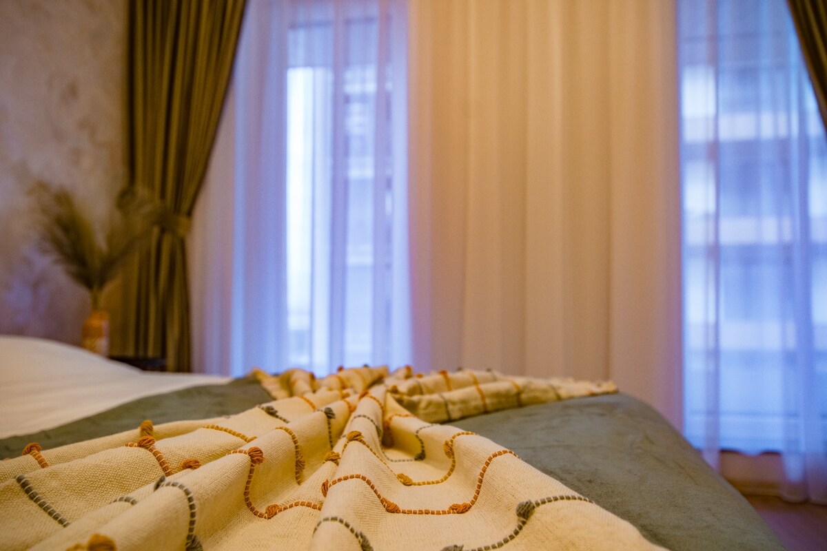 A cozy bedroom is depicted, featuring soft curtains that frame a window, allowing diffused light to fill the space. A textured blanket is casually arranged over a spacious bed, adding a touch of warmth to the inviting atmosphere.