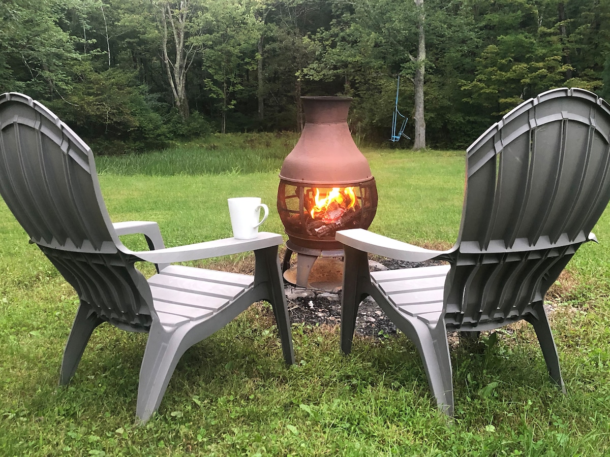 Two gray patio chairs are positioned around a fire pit, which is adorned with flickering flames. A white mug is placed on a nearby surface, while a grassy area and leafy trees provide a serene backdrop.