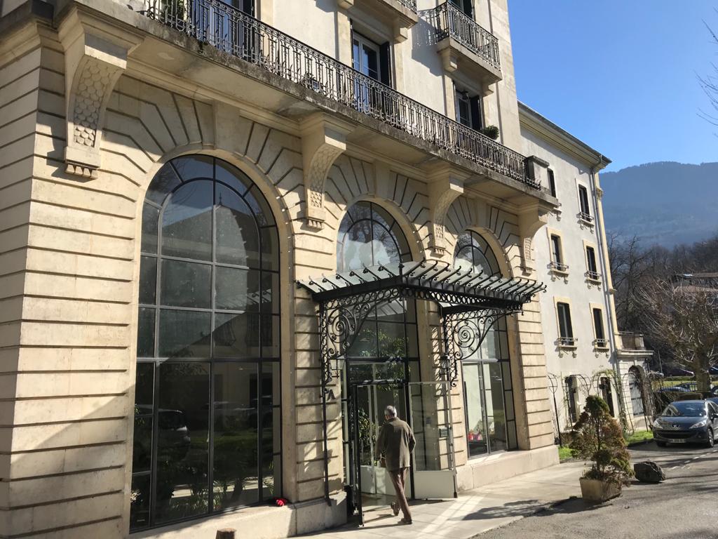 The elegant exterior of a historic building is highlighted by large arched windows and decorative balconies. The entrance features a beautifully designed awning supported by ornate ironwork. The surrounding landscape includes greenery and mountains in the background under clear blue skies.