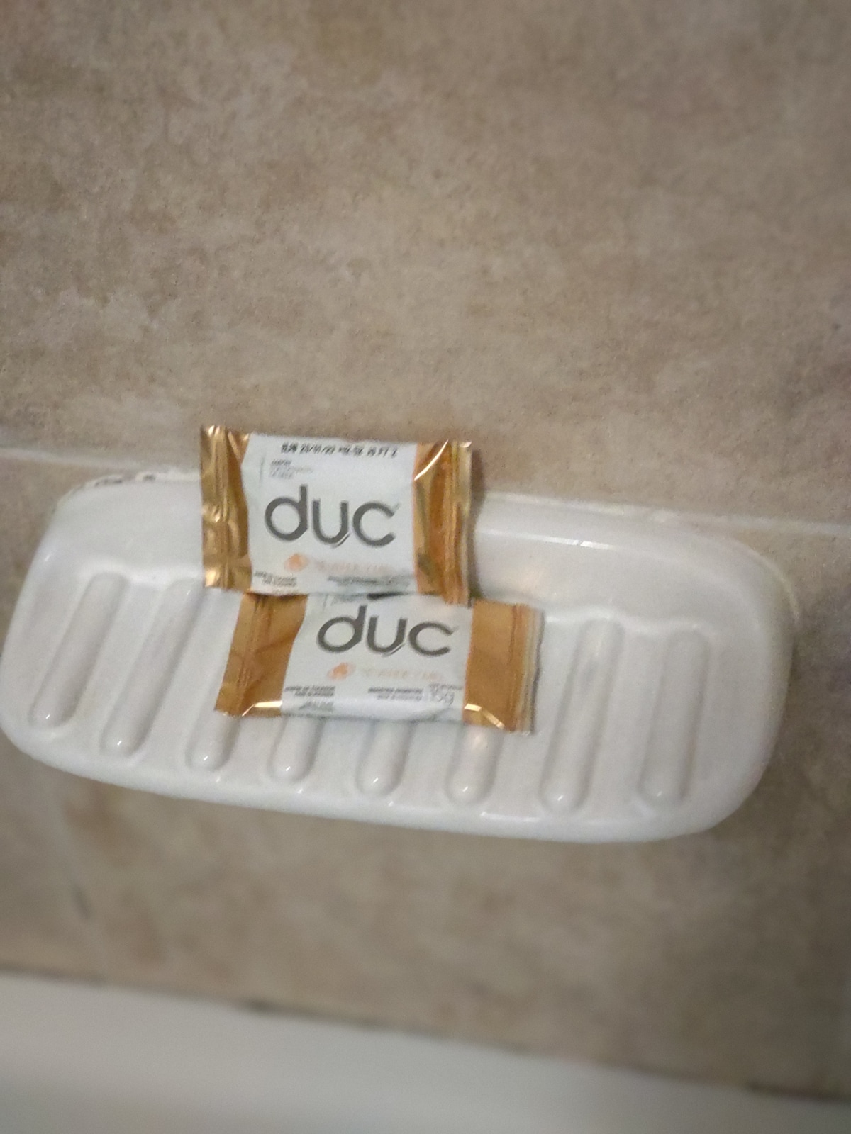 Two bars of soap are presented on a white, textured soap dish against a beige tiled wall. The packaging features gold and white colors with the word 'duc' prominently displayed.