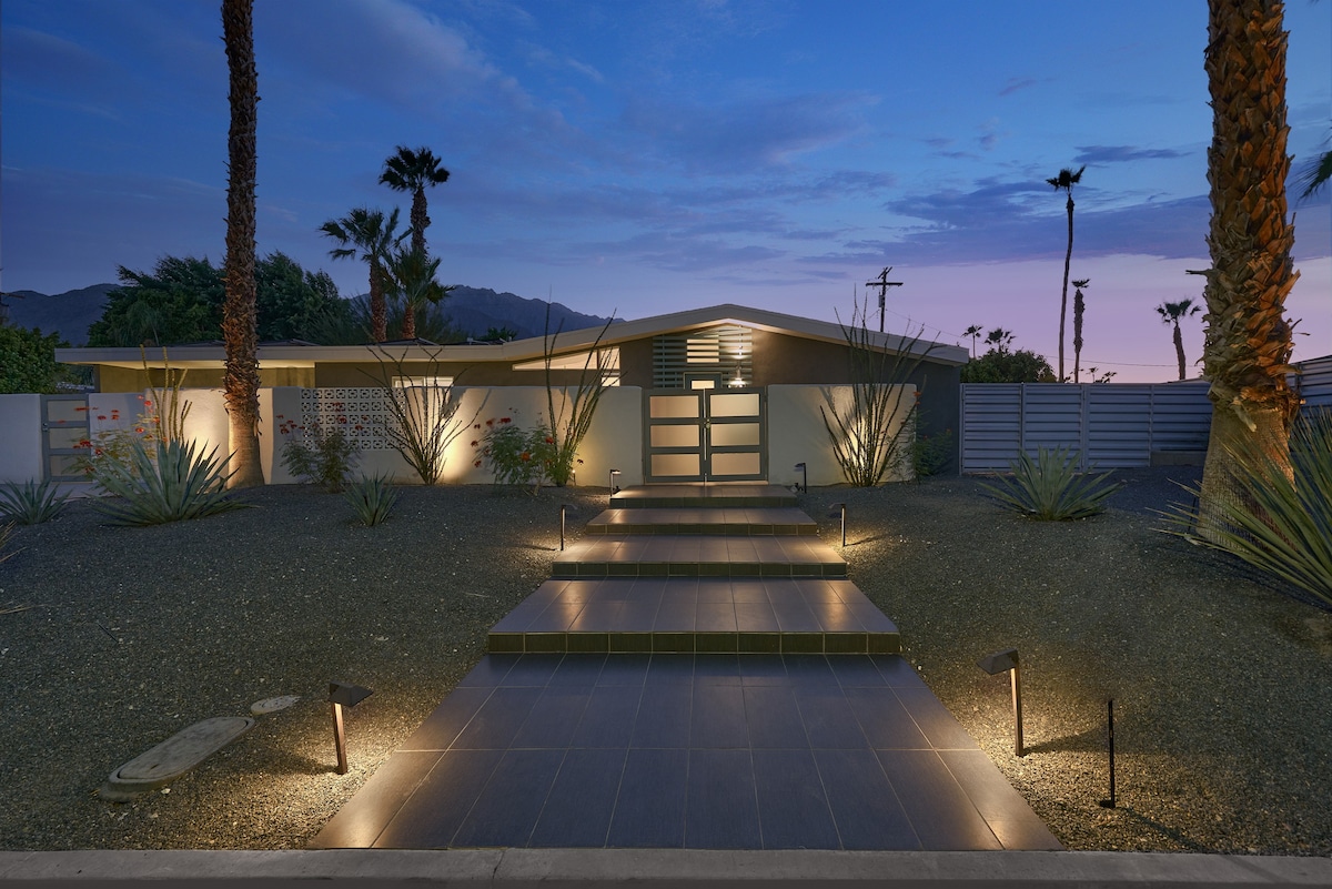 The entrance to the mid-century home is illuminated by soft landscape lighting, highlighting a pathway lined with modern stepping stones. Surrounding greenery, including agave plants and palm trees, enhances the serene atmosphere, with a clear evening sky in the background.