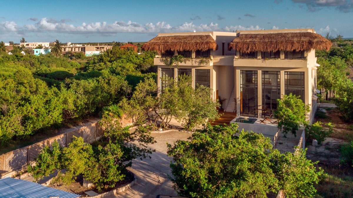 The exterior of Casa Imox is showcased, featuring a modern structure with large glass windows and a thatched roof. Surrounding greenery adds to the natural setting, while the distant landscape offers glimpses of nearby buildings and open sky.