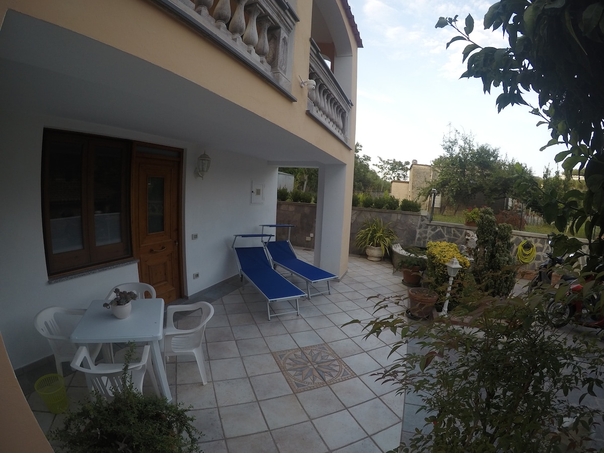 An outdoor terrace features two blue lounge chairs positioned beside a small white table. Potted plants are arranged around the space, contributing greenery. A stone path leads to the entrance, while the building's exterior displays a light-colored facade and ornamental balcony.