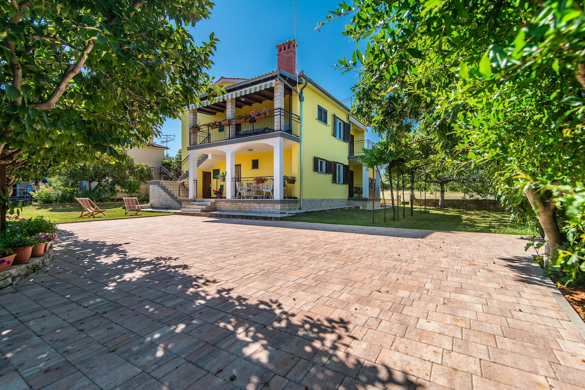 A bright yellow house features a welcoming two-story design, with a spacious terrace adorned with seating. The brick-paved yard is framed by lush greenery and flowering plants, providing an inviting outdoor space for relaxation.