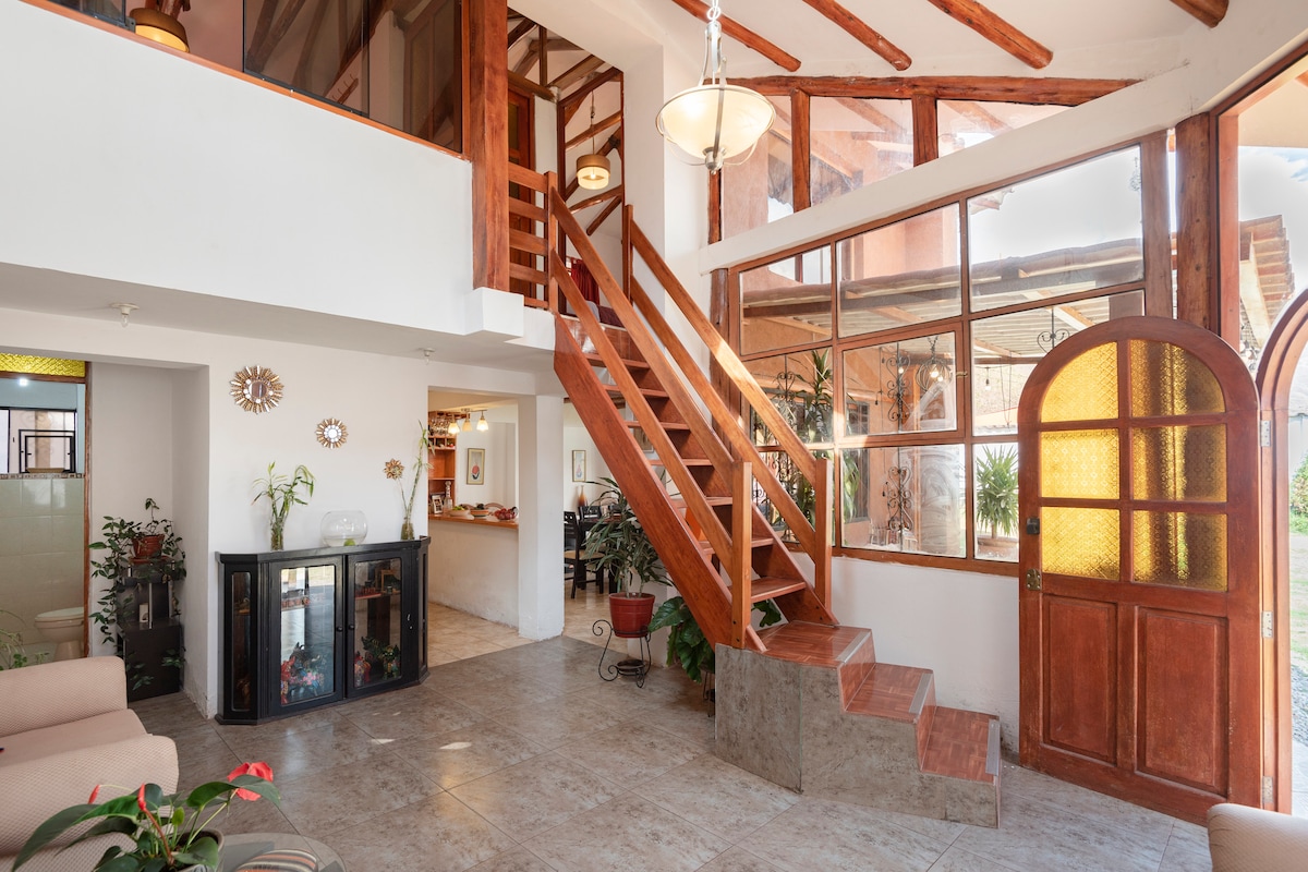 The entryway features an open, two-story layout with large windows allowing natural light to enhance the space. A wooden staircase leads to the upper level, while a cozy seating area is visible to the left, framed by neutral colors and indoor plants.