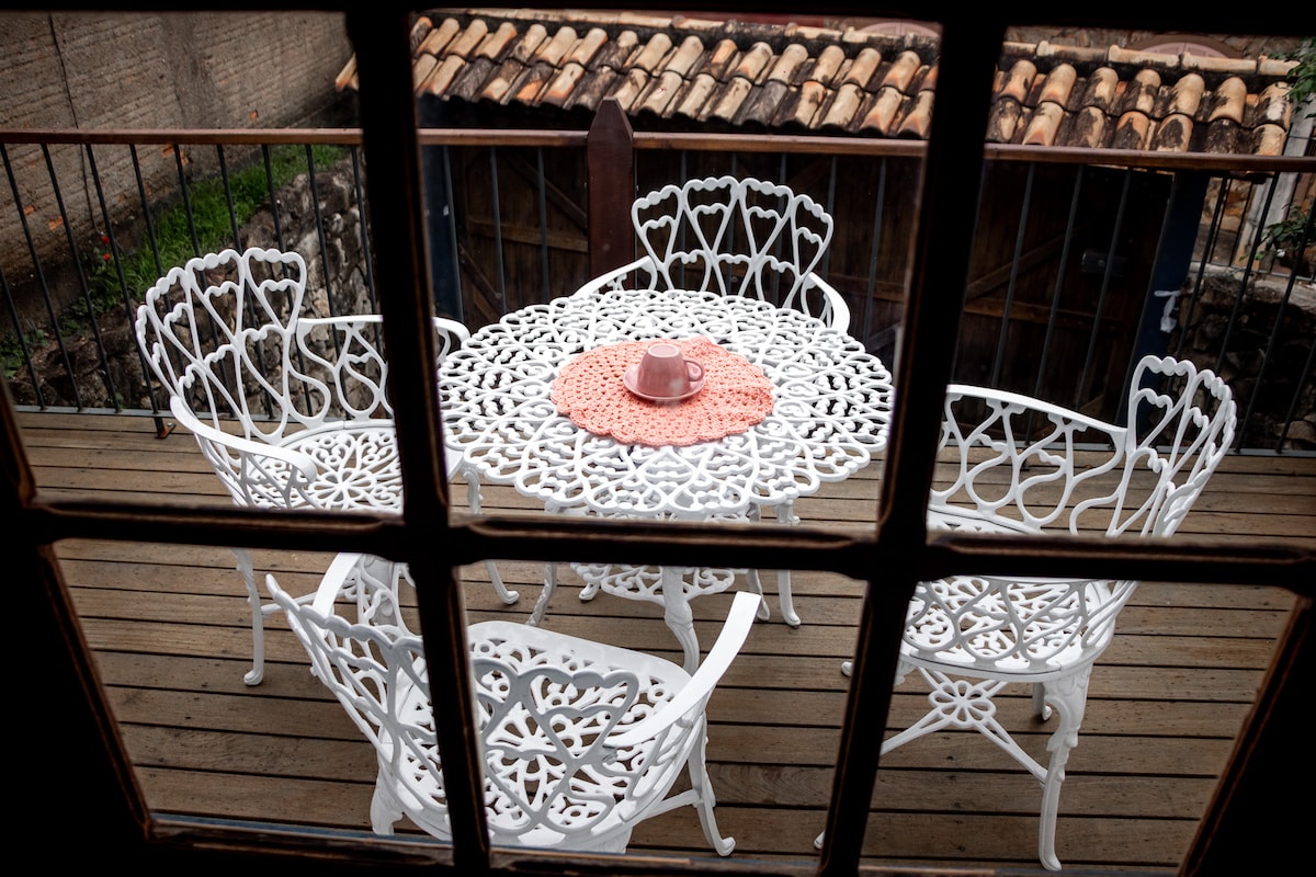 A charming outdoor deck area is visible through the window, featuring a white wrought-iron table surrounded by four matching chairs. A circular placemat and a cup are placed on the table, evoking a space for relaxation and outdoor dining.