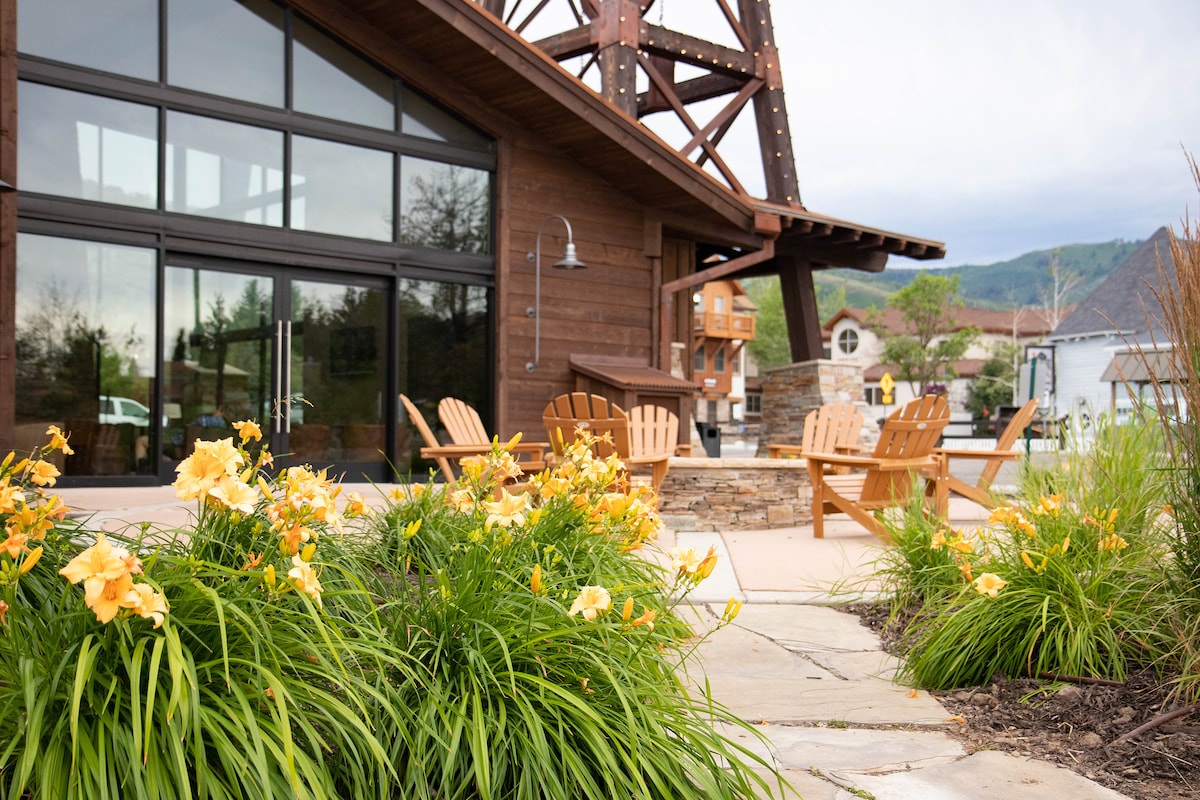 A welcoming outdoor area is surrounded by vibrant greenery and blooming yellow flowers. Several wooden chairs are positioned on a stone patio, offering a comfortable space for relaxation. The modern building features large glass windows that enhance the connection with the surrounding landscape.