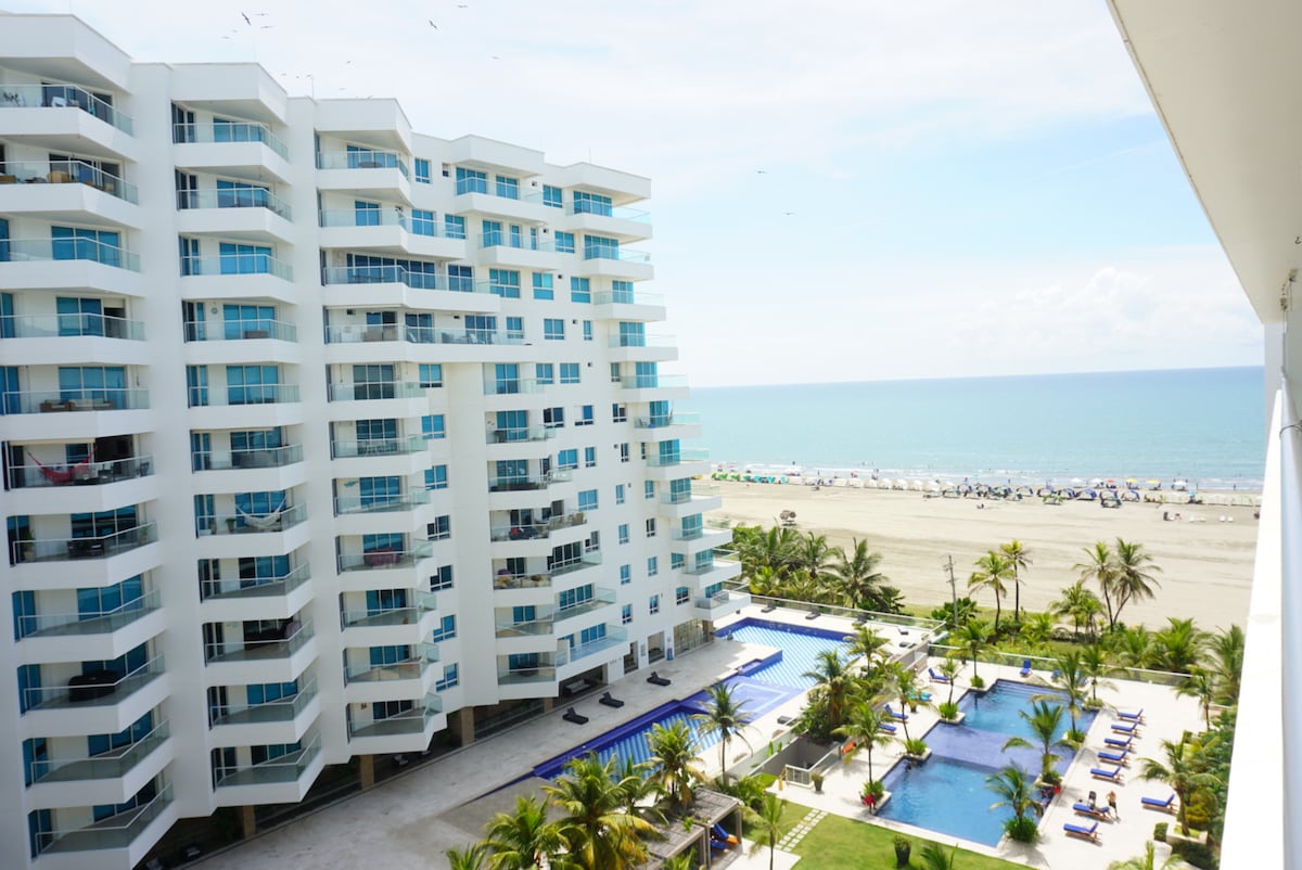 A panoramic view from the apartment balcony reveals the ocean alongside a sandy beach. An outdoor pool area is visible, surrounded by palm trees and lounge chairs. The building's modern architecture features multiple balconies with glass railings, highlighting the inviting atmosphere of the space.