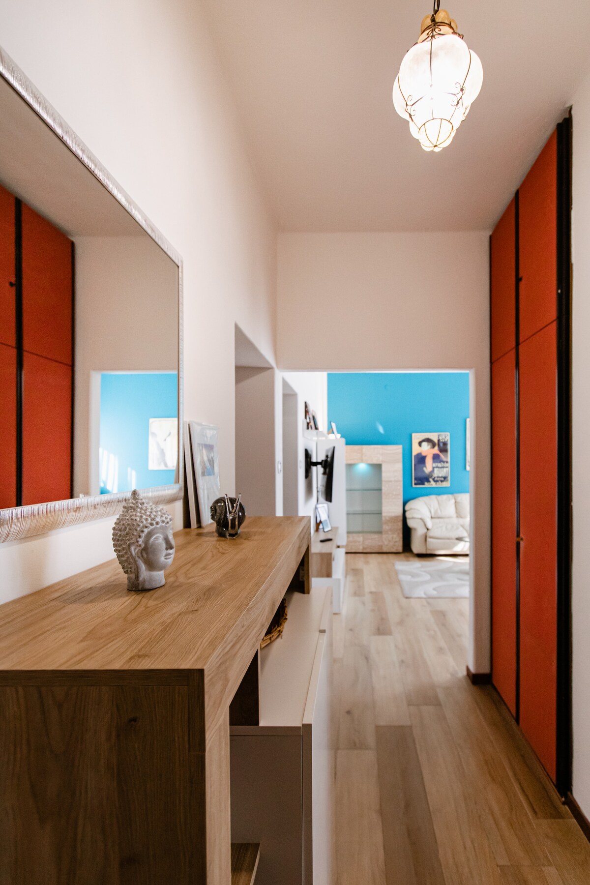 A hallway is displayed with a light wooden console and a decorative statue. The walls are accented with vibrant blue and red panels. A mirror adorns the console, while a softly glowing pendant light hangs above, illuminating the space. A glimpse of a cozy living area is visible at the end.