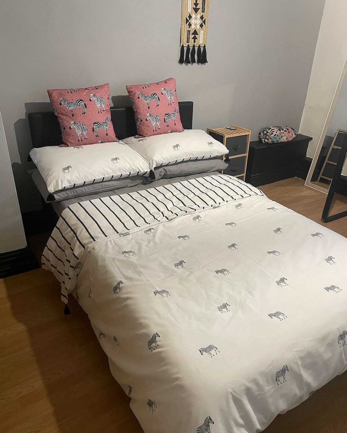 A small double bed is paired with decorative pillows featuring a zebra pattern. The bedding includes a playful design of zebras, complemented by a striped blanket. A side table and a patterned bag are visible in the corner, adding functionality to the cozy room.