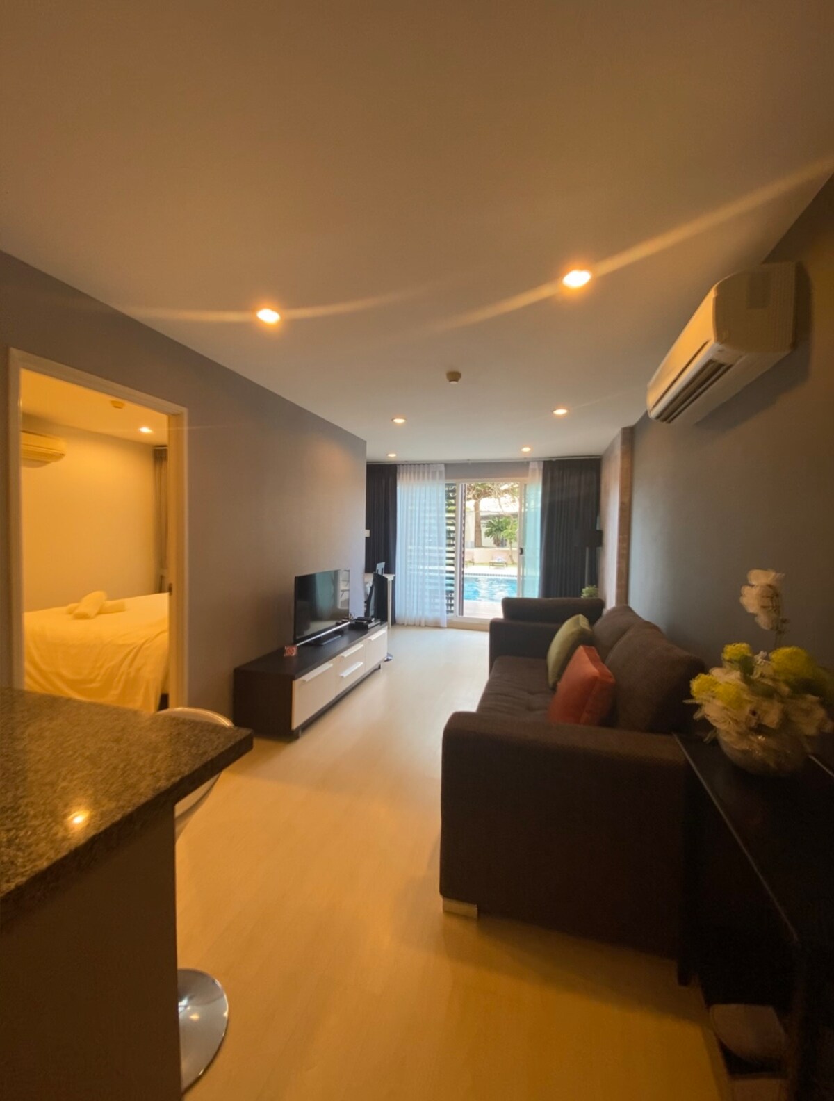 A well-lit living area features a comfortable sofa with colorful cushions and a flat-screen television. Light from a sliding glass door illuminates the space, leading to an inviting pool area. A small dining table is visible near the kitchen counter, enhancing functionality.