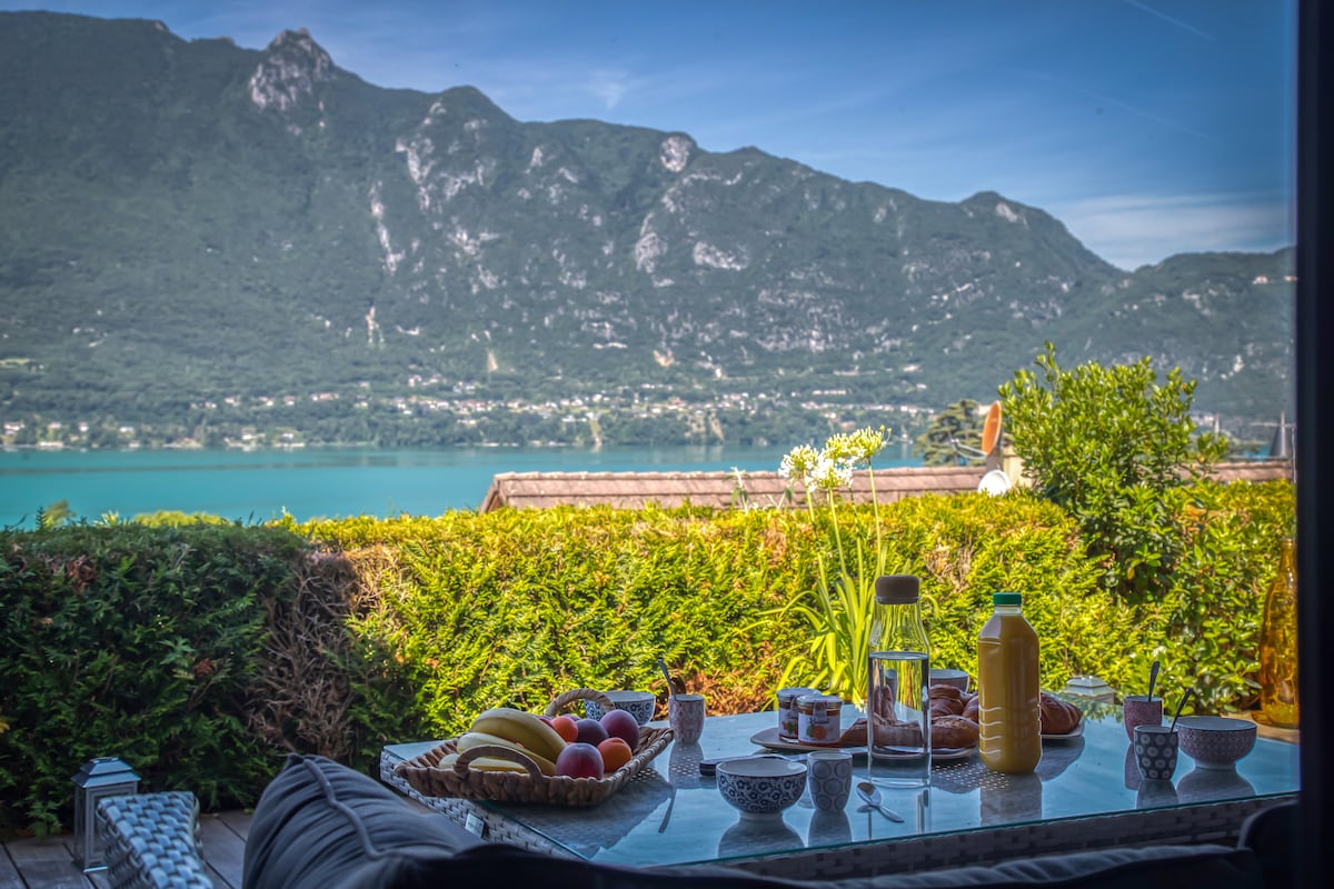 A dining table is set on a patio, adorned with fresh fruits and pastries. Glass bottles and dishes complement the serene view of the lake and mountains in the background, framed by lush greenery. The scene conveys a peaceful outdoor dining experience.