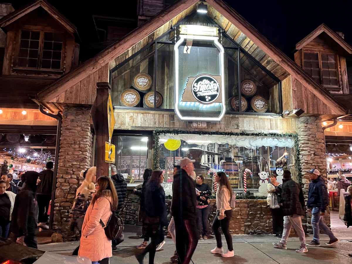 A lively scene outside a charming store, featuring wooden architecture and a glowing neon sign. People stroll by, engaged in conversation. The storefront is decorated for the evening, with festive lights enhancing the inviting atmosphere during a bustling night.