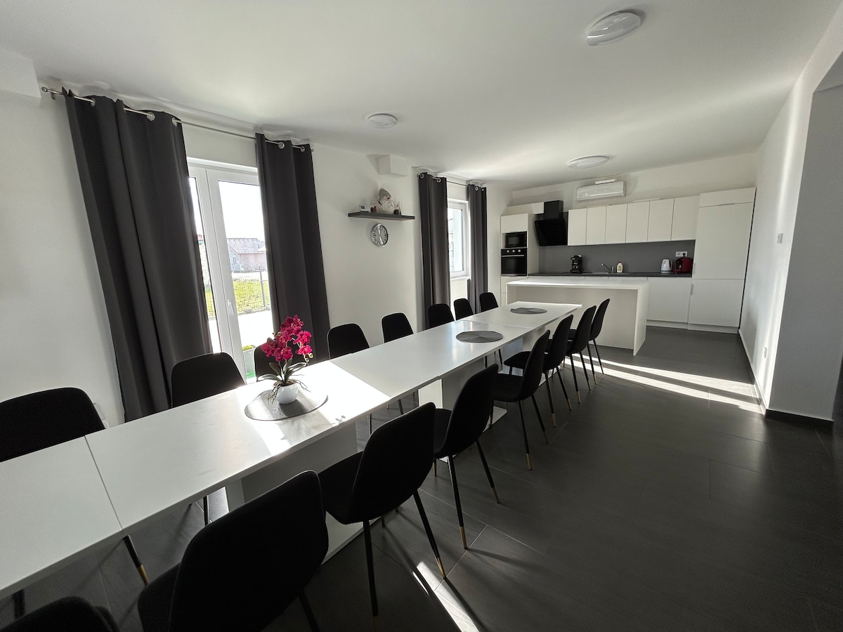 A spacious dining area features a long white table surrounded by twelve black chairs. Natural light saturates the room through large windows, enhancing the modern kitchen equipped with sleek appliances. Dark curtains frame the windows, adding a touch of elegance to the bright space.