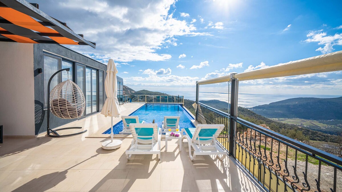 A stunning outdoor area is presented with a sparkling swimming pool surrounded by lounge chairs. A soft, white umbrella provides shade, while a hanging chair adds a relaxed touch. The panoramic view of the mountains and sea is highlighted under a bright blue sky with soft clouds.