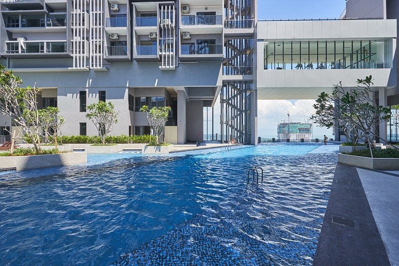 An expansive infinity swimming pool is showcased, reflecting the sky. Surrounding trees and greenery are visible along the pool's edges, providing a serene atmosphere. The modern condominium structure is partially seen in the background, featuring large glass windows that enhance the openness.
