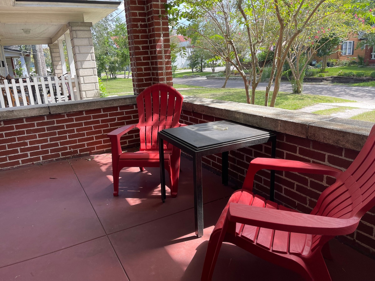 A covered sitting porch is depicted, featuring two red chairs and a small black table. The floor is tiled in a warm tone, with greenery visible beyond the brick walls, offering a peaceful view of the surrounding area.