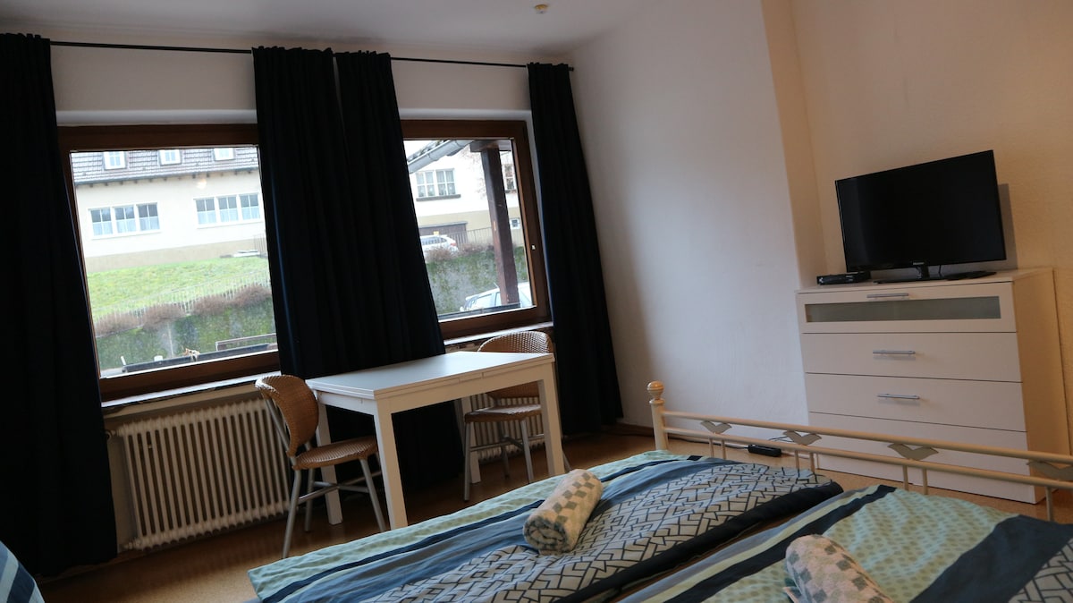 A sunny bedroom features large windows with dark curtains overlooking a grassy area. A spacious bed with patterned bedding is placed beside a sleek white dresser. A small table with two chairs creates a functional space, while a television is mounted on the wall.