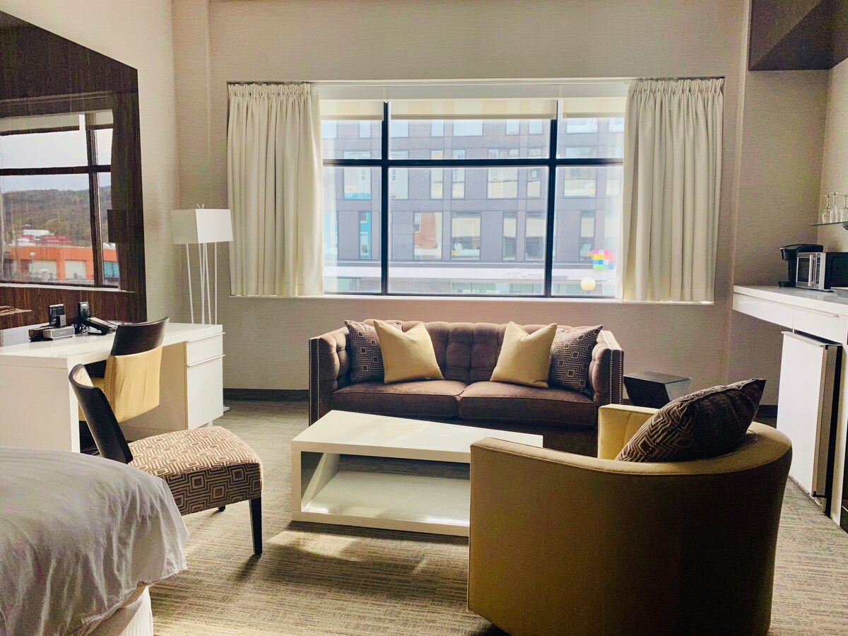 An inviting living space features large windows allowing ample natural light. A comfortable brown sofa is centered with accent pillows, accompanied by two armchairs. A stylish coffee table rests in front while a dresser and workspace are visible, providing an optimal area for relaxation.