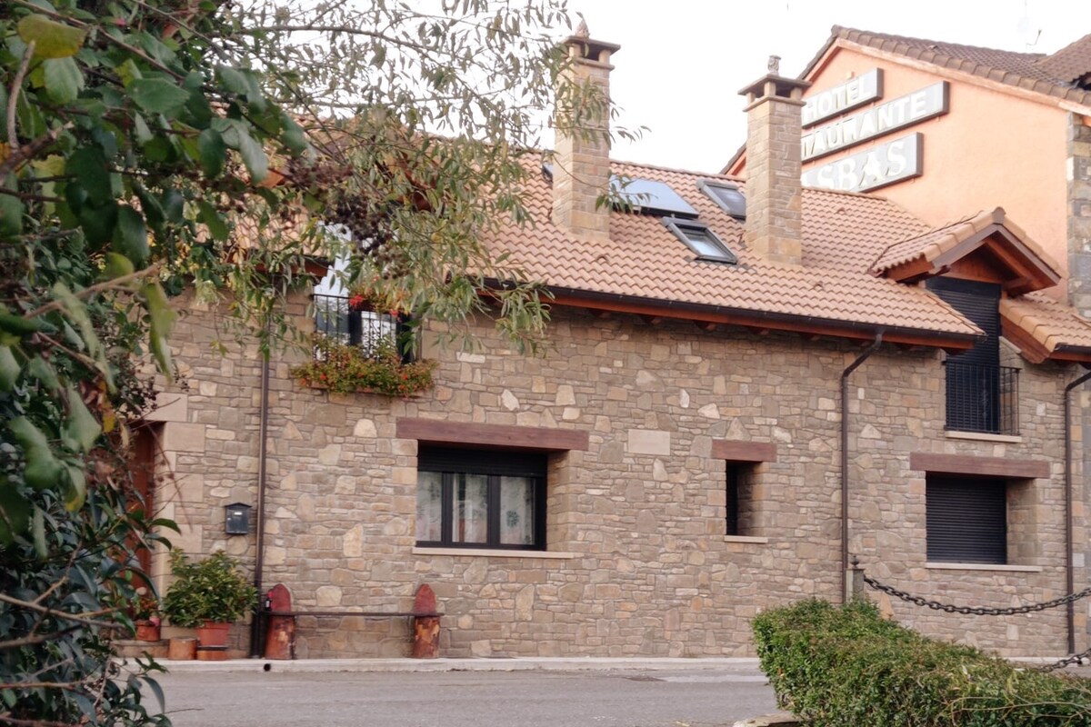 A traditional stone facade characterizes the building, featuring multiple levels and a tiled roof. Flower boxes are positioned on the upper window, adding a touch of greenery. The entrance is framed by a small stone wall and potted plants, contributing to the welcoming exterior.