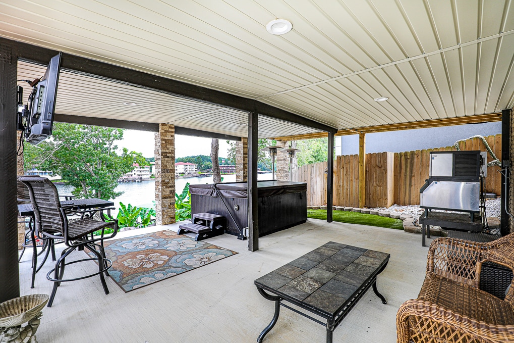 A covered outdoor area features comfortable seating and a coffee table, providing a relaxing space. A hot tub overlooks the lake, while a television is mounted nearby. A grill is positioned on one side, and greenery adds a touch of nature to the setting.