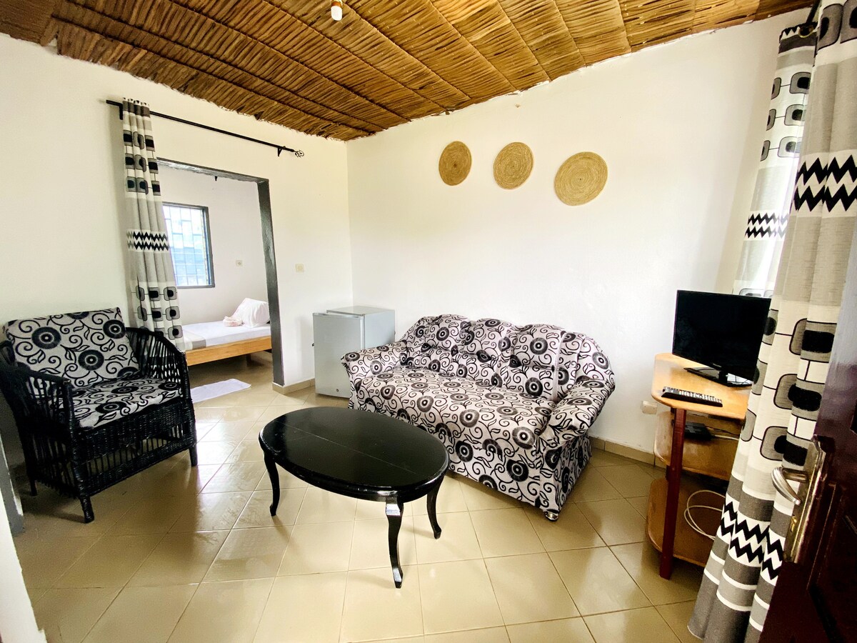 A comfortable seating area features a patterned sofa and two chairs, complemented by a round coffee table. A small refrigerator and a television are visible, with light flowing through window treatments that add a touch of style to the warm, inviting space.